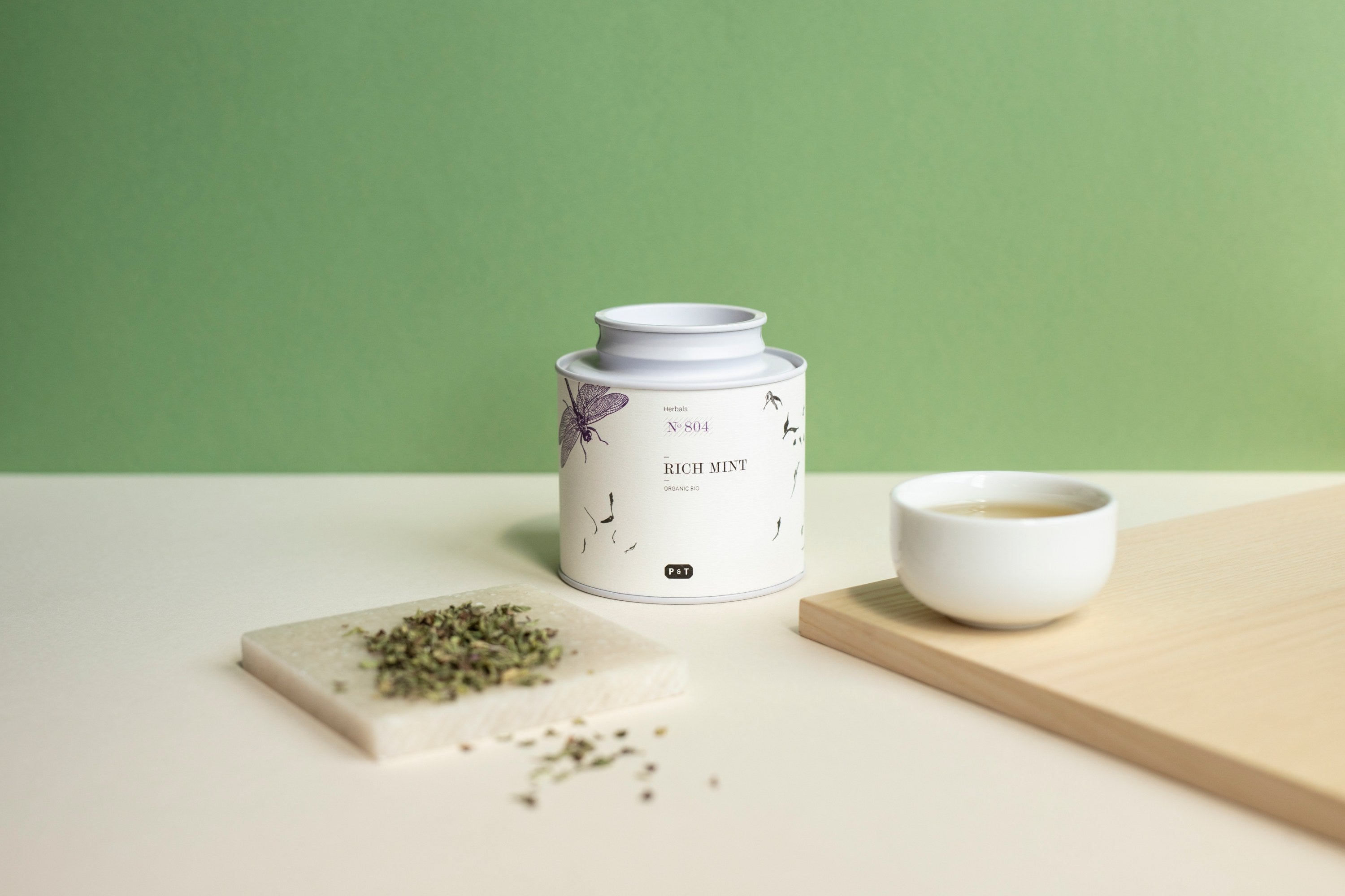 Weiße Teedose mit der Aufschrift ‚RICH MINT‘ und Illustrationen von Blättern und Schmetterlingen, daneben eine kleine weiße Schale mit Tee und eine quadratische Marmorplatte mit getrockneten Minzblättern vor grünem Hintergrund