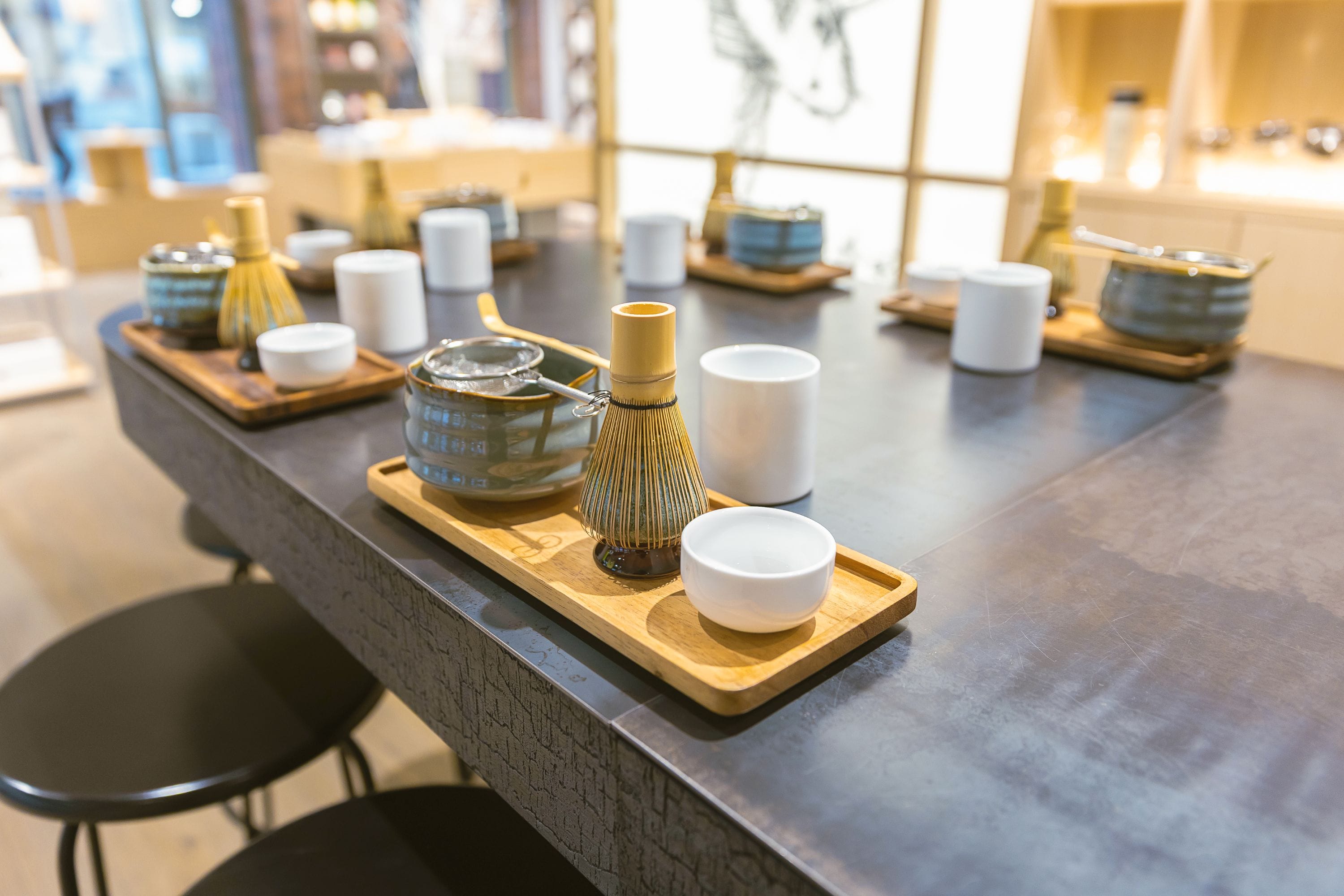 Moderner Teetisch mit mehreren Holztabletts, jeweils mit Teekanne, Besen und Tassen – umgeben von Hockern, im Hintergrund Regale mit verschiedenen Gegenständen