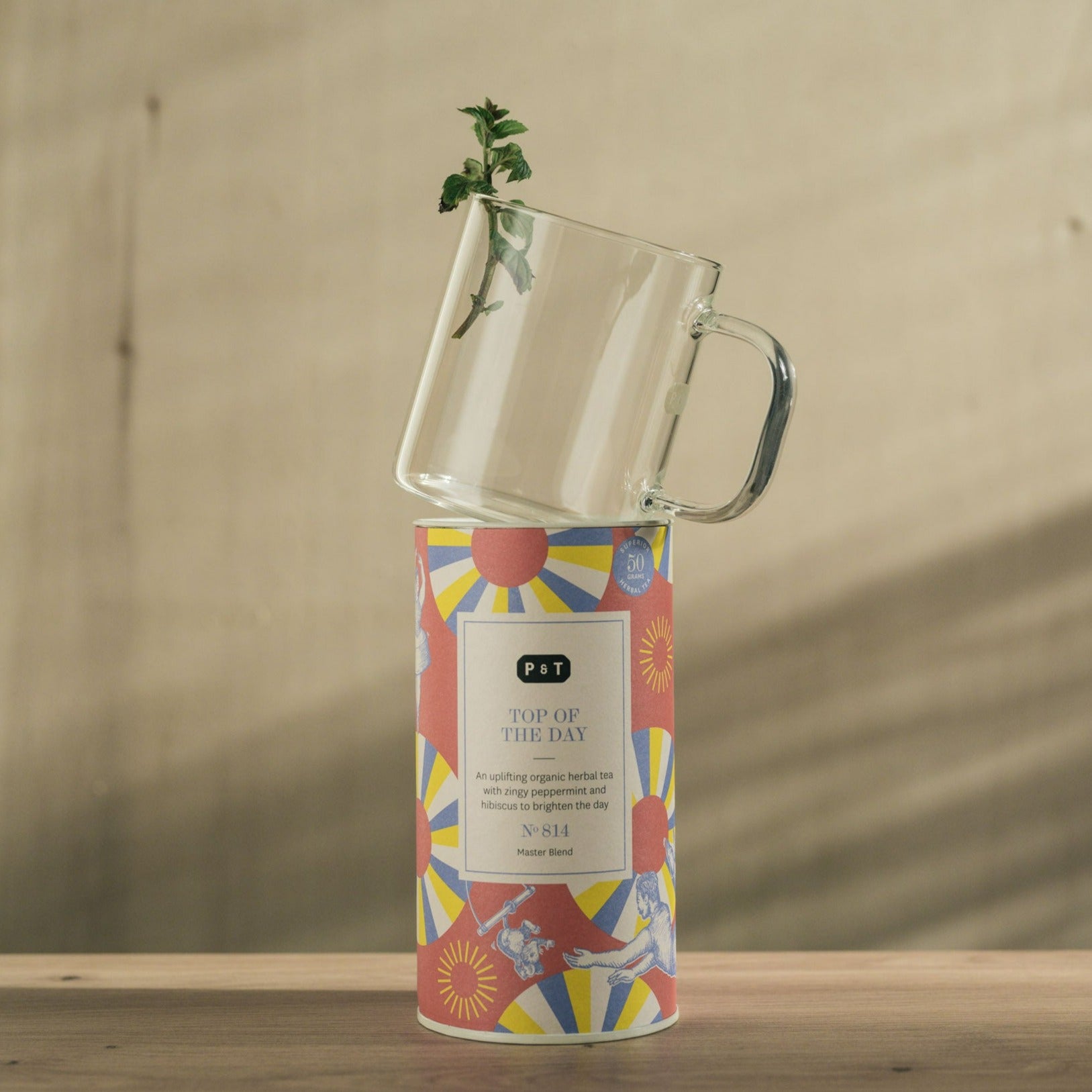 Zylindrische P & T Teedose mit sonnigem Muster neben Glas mit frischer Pfefferminze auf Holz vor beigem Hintergrund