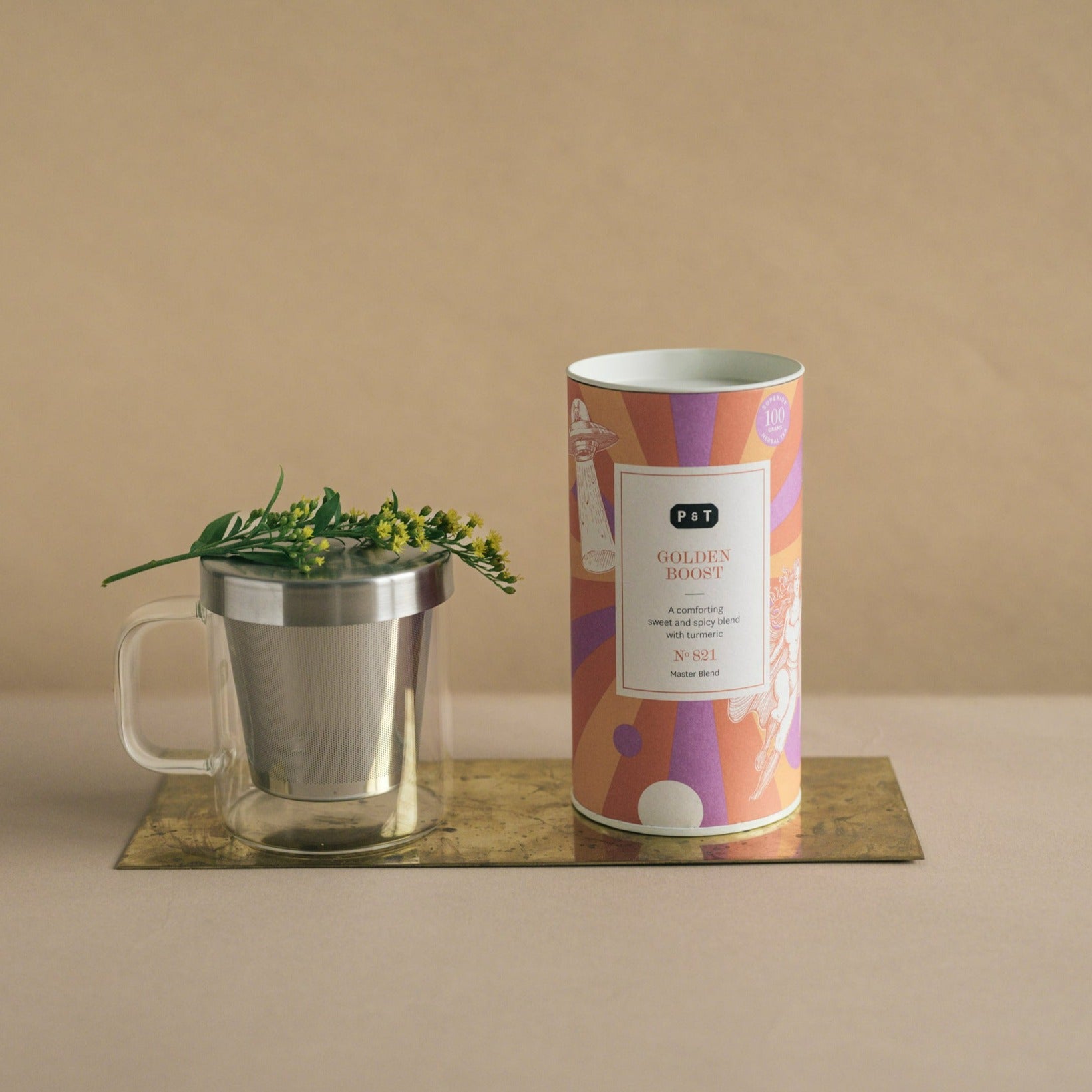 Zylindrische Teeverpackung P & T GOLDEN BOOST mit Kurkuma-Geschmack neben Glasbecher mit Metalldeckel.