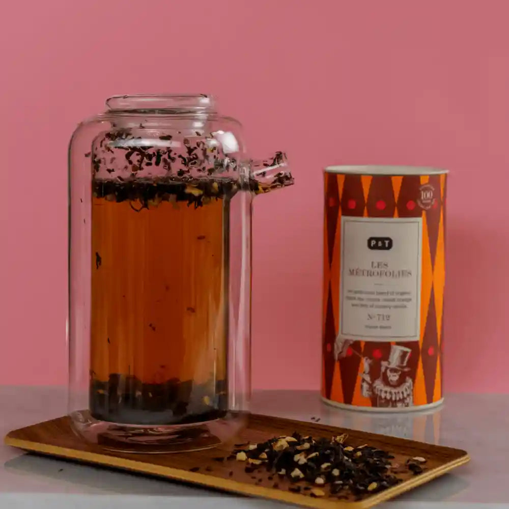 Glaskanne mit bernsteinfarbenem Tee und losen Blättern neben einer P & T Les Metropolies Teedose.