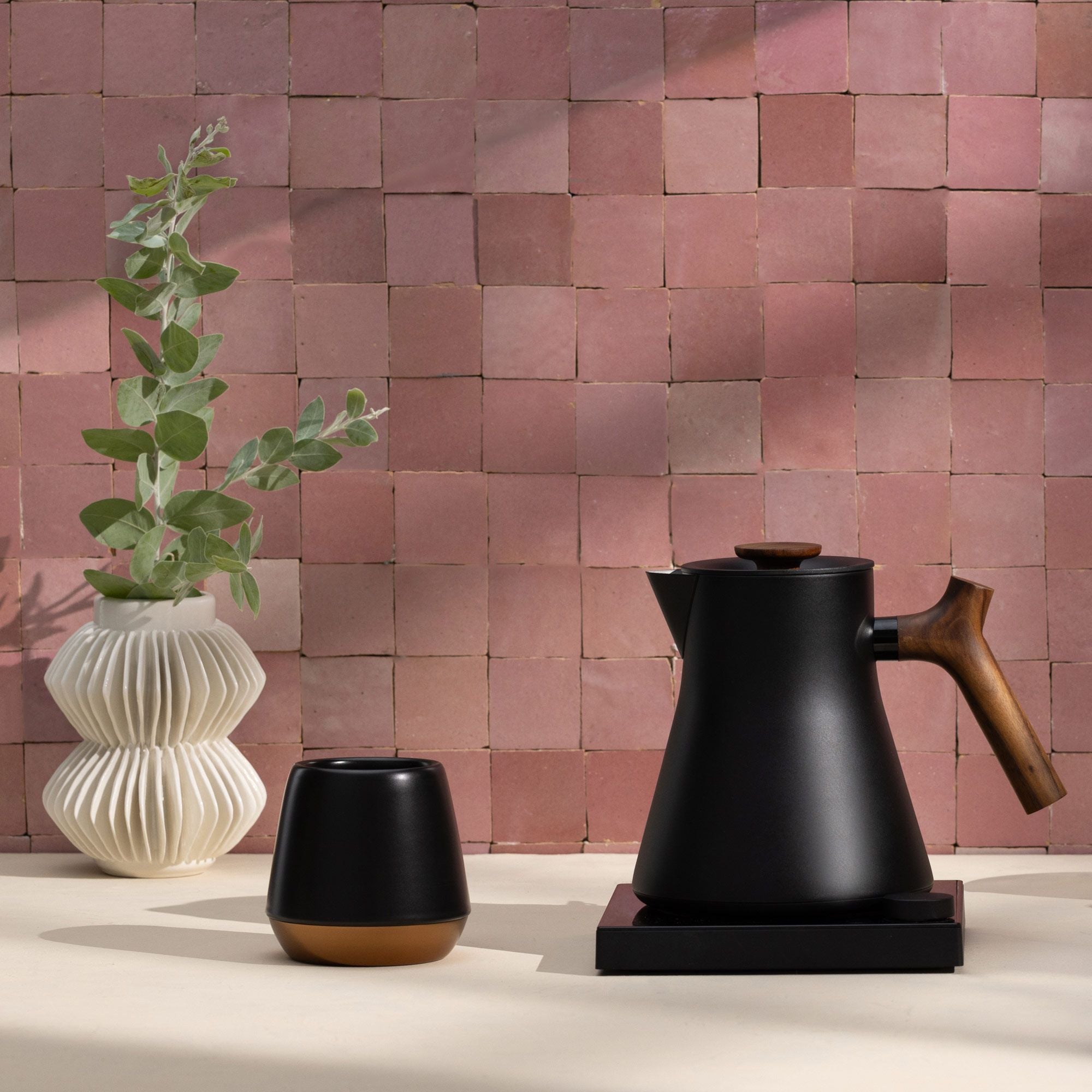 Mattierter schwarzer Wasserkocher mit Holzgriff, kleine Tasse neben cremefarbener Vase und rosarotem Hintergrund.
