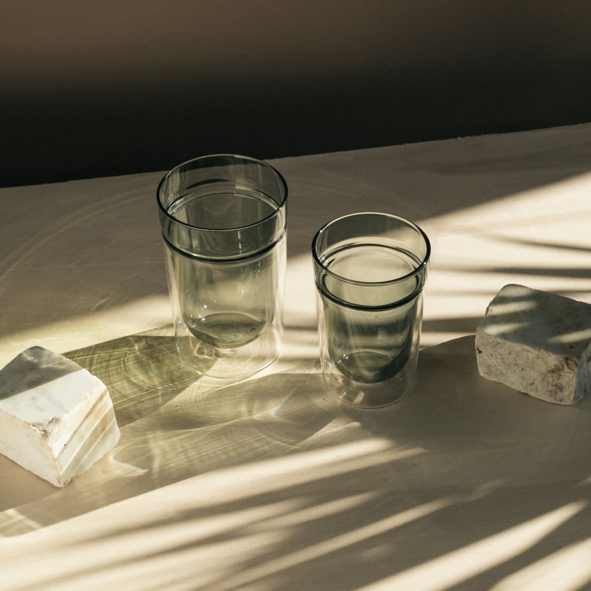 Zwei transparente Trinkgläser in unterschiedlichen Größen auf beiger Oberfläche mit Sonnenlicht.