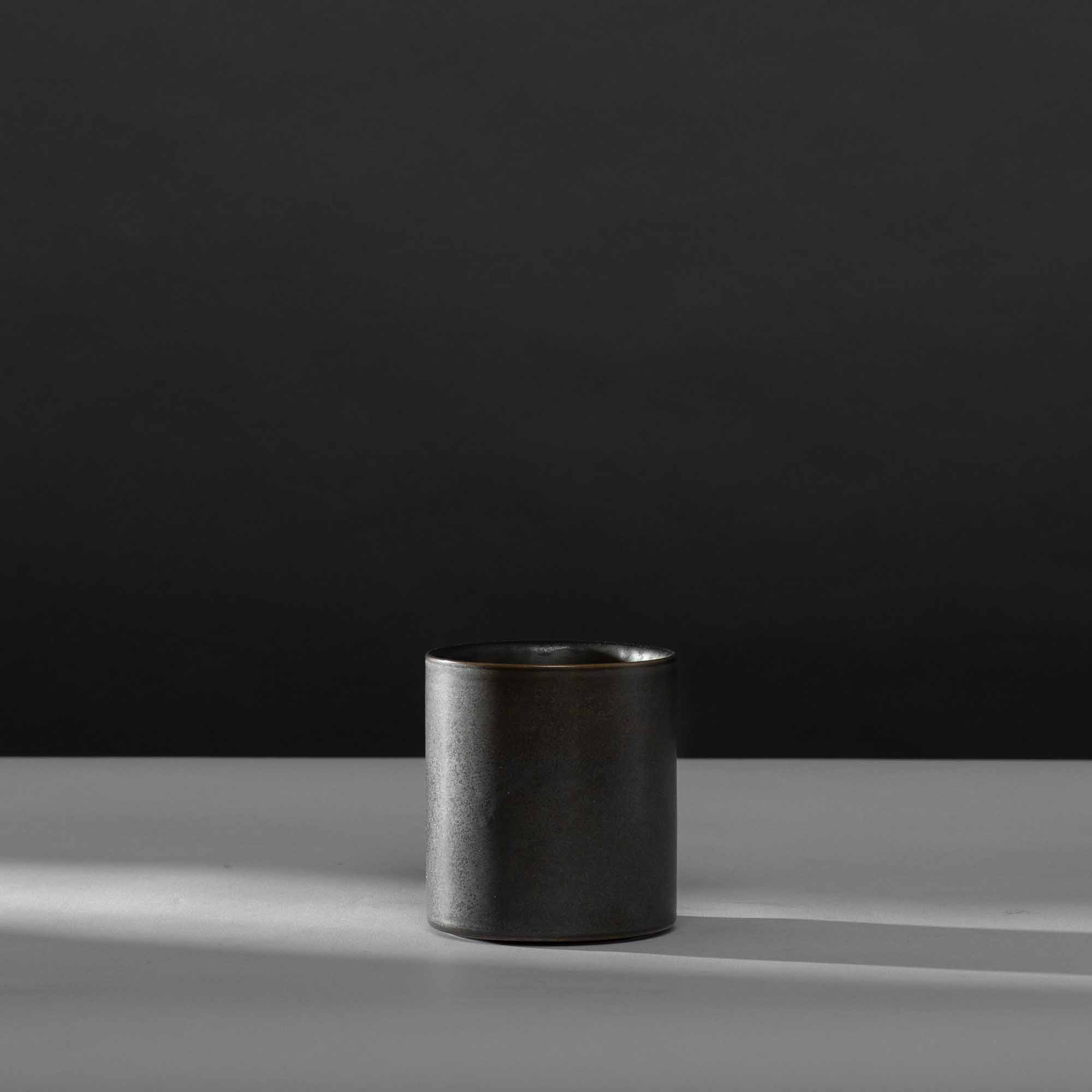 Matte schwarze Keramiktasse auf grauem Tisch vor schwarzem Hintergrund.