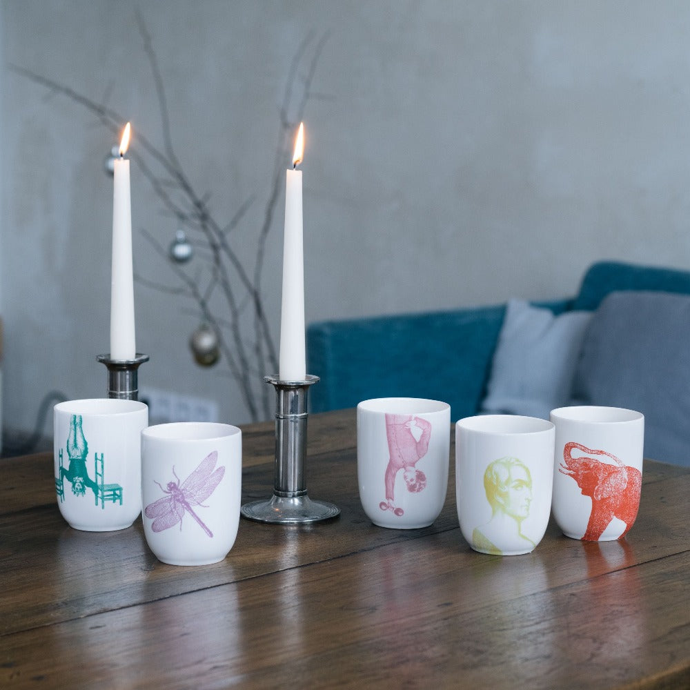 Fünf weiße Keramiktassen mit bunten Mustern und zwei brennende weiße Kerzen in silbernen Haltern auf einem Holztisch.
