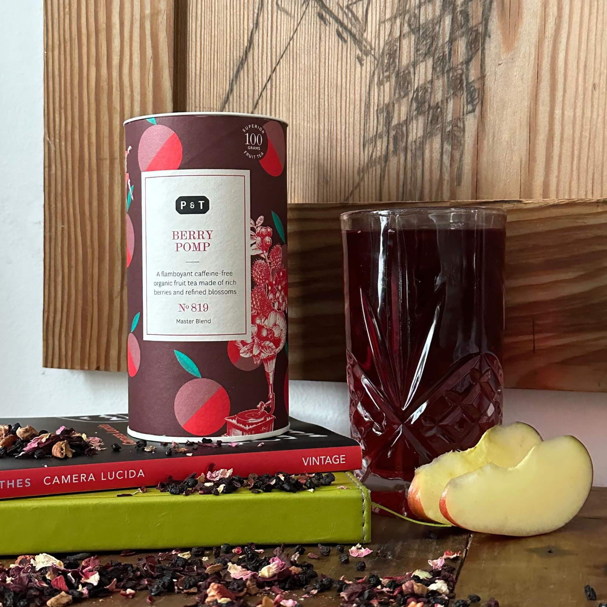 Zylindrische Dose Bio-Beerentee 'Berry Pomp', Glas mit rotem Früchtetee, Pfirsichscheiben und Vintage-Bücher auf Holz.