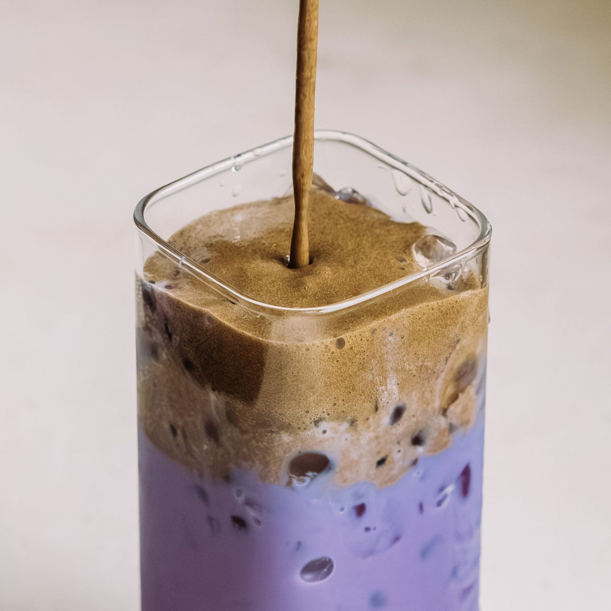 Glas mit violettem Eistee und schaumiger brauner Haube, mit Eiswürfeln, vor hellem Hintergrund