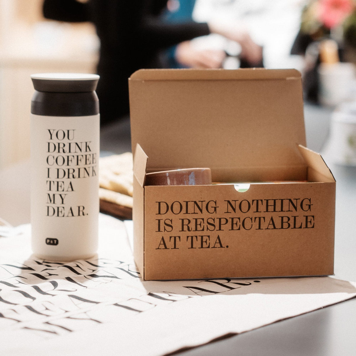 Weißer Reisebecher mit Aufdruck 'You drink coffee, I drink tea my dear' neben geöffneter brauner Box.