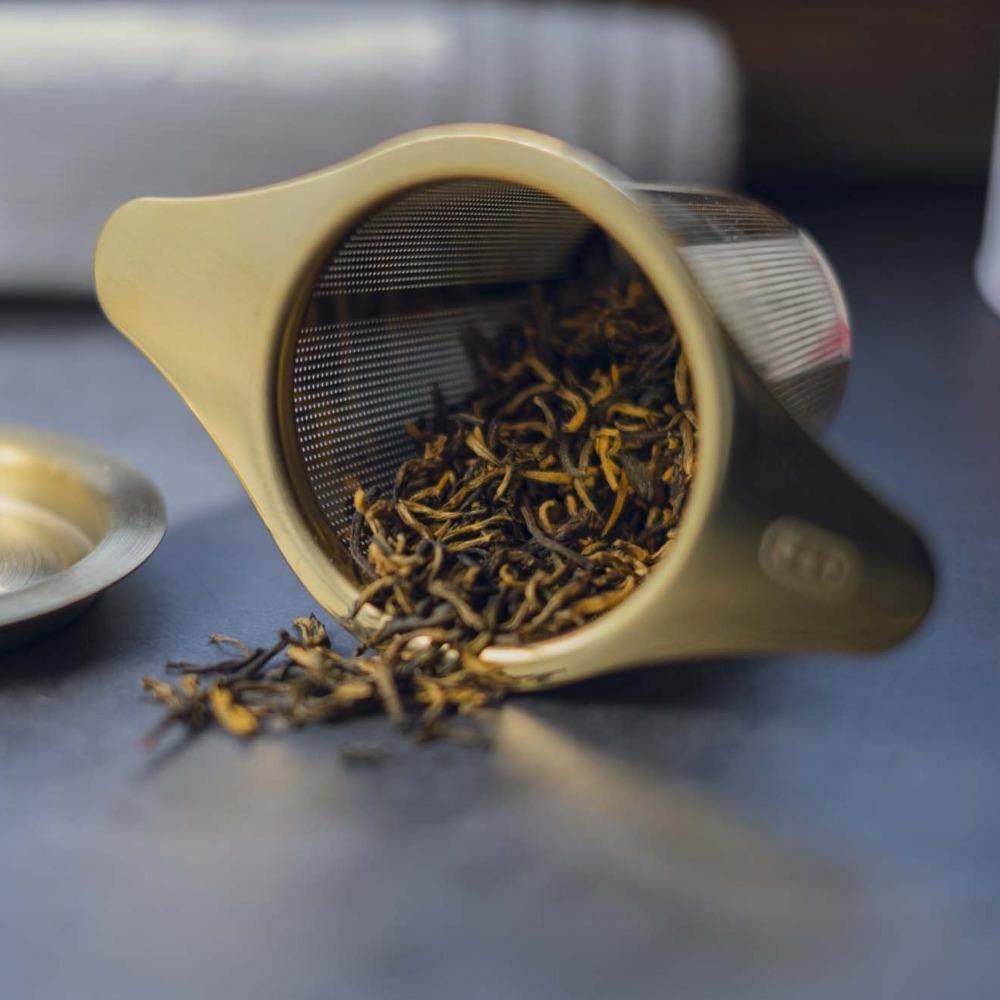 Messing-Teesieb mit losem Tee, seitlich liegend, mit CGK-Schriftzug und sanfter Beleuchtung.