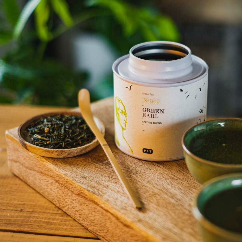 Weißer Behälter mit 'GREEN EARL' Tee und P & T Logo, lose Teeblätter, Holzlöffel, zwei Keramiktassen auf Holztisch.