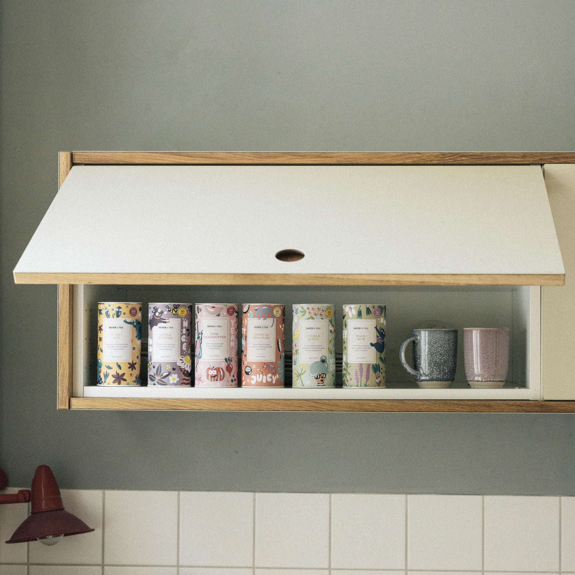 Sechs bunte Teedosen mit Designs und Schriftzügen in weißem Küchenregal neben zwei Keramiktassen.