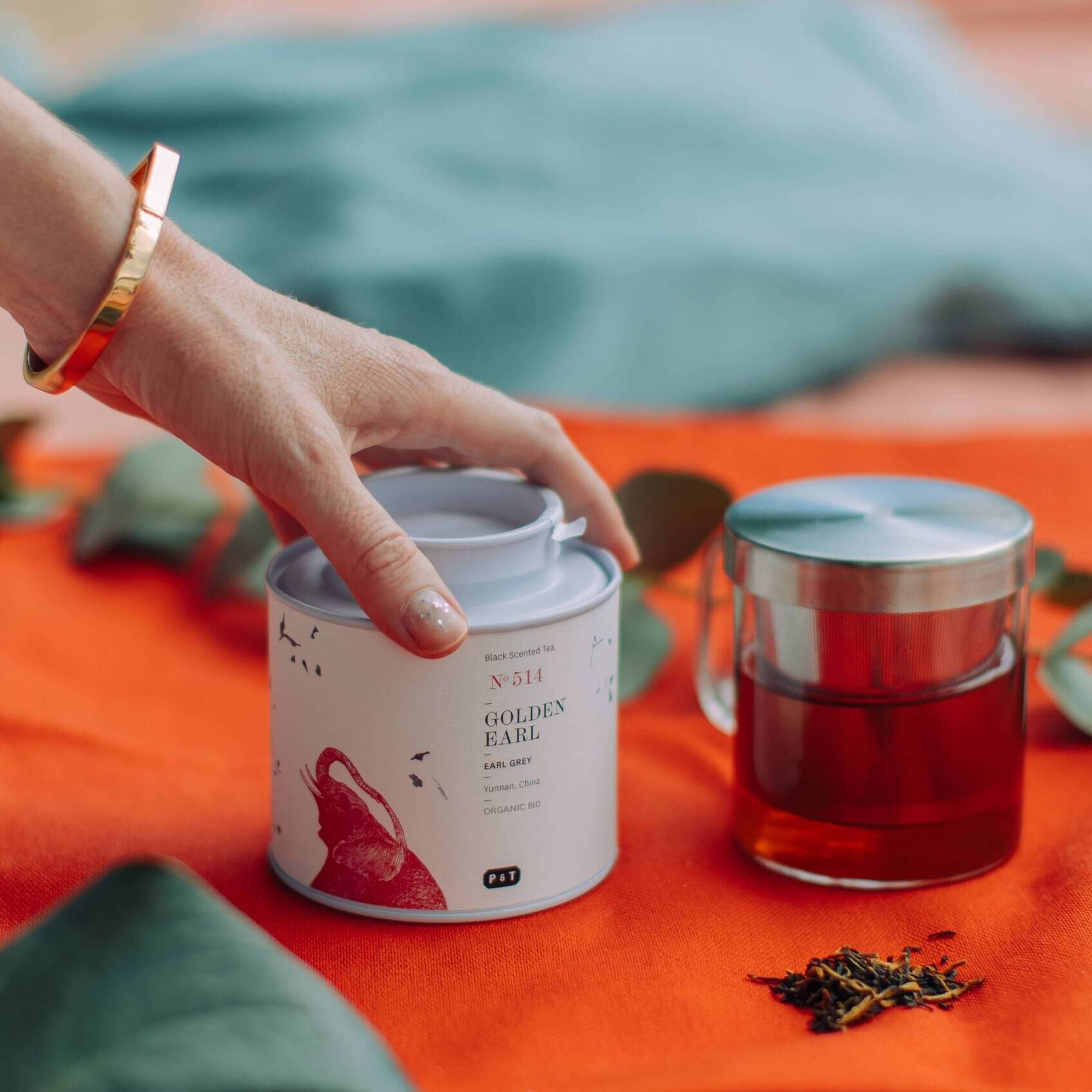 Hand öffnet weiße GOLDEN EARL Teedose neben Glas-Teekanne mit dunklem Earl Grey Tee und losen Blättern.