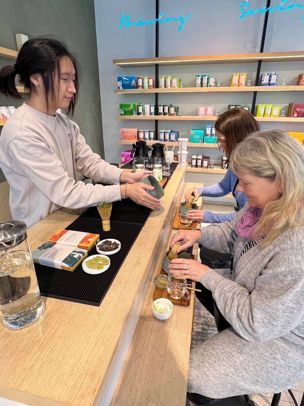 Drei Personen bei einer Teezeremonie an einer Holztheke mit Bambusbesen und Keramikschalen, umgeben von bunten Teepackungen.