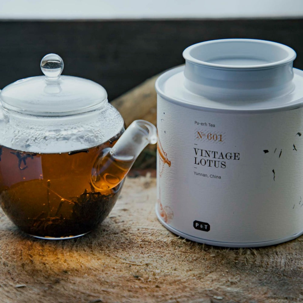 Glaskanne mit bernsteinfarbenem Pu-erh-Tee, Teeblätter daneben, und weiße Dose mit Vintage Lotus Tee aus Yunnan.
