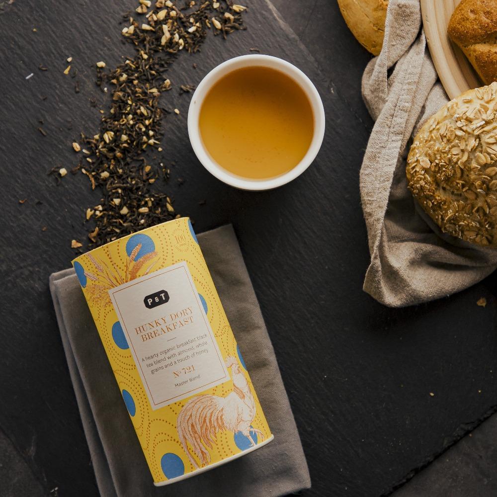 Gelbe Teedose 'HUNKY DORY BREAKFAST' mit Hahn und blauen Kreisen, weiße Tasse mit Tee, Brötchen auf beigem Tuch.