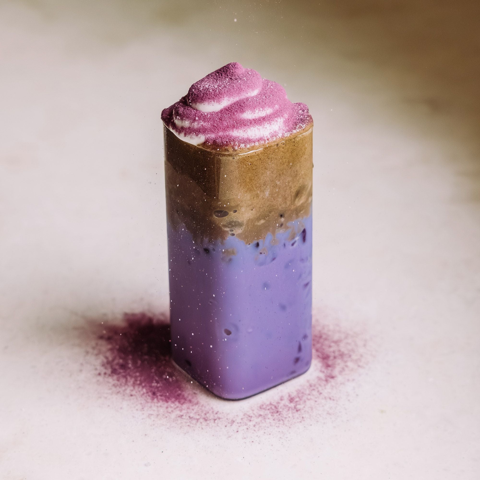 Dreischichtiges Getränk in hohem Glas mit lila Eis, Kaffee-Textur und cremigem Schaum auf beige Oberfläche
