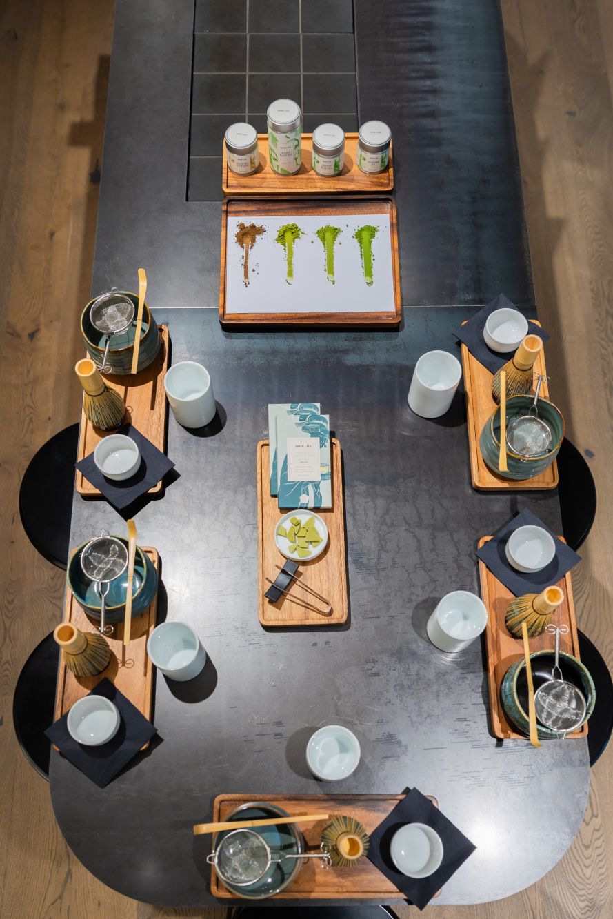 Schwarzer ovaler Tisch mit Holztabletts, Keramiktassen, Bambus-Teewerkzeugen und vier Tee-Jars mit Matcha-Sorten.