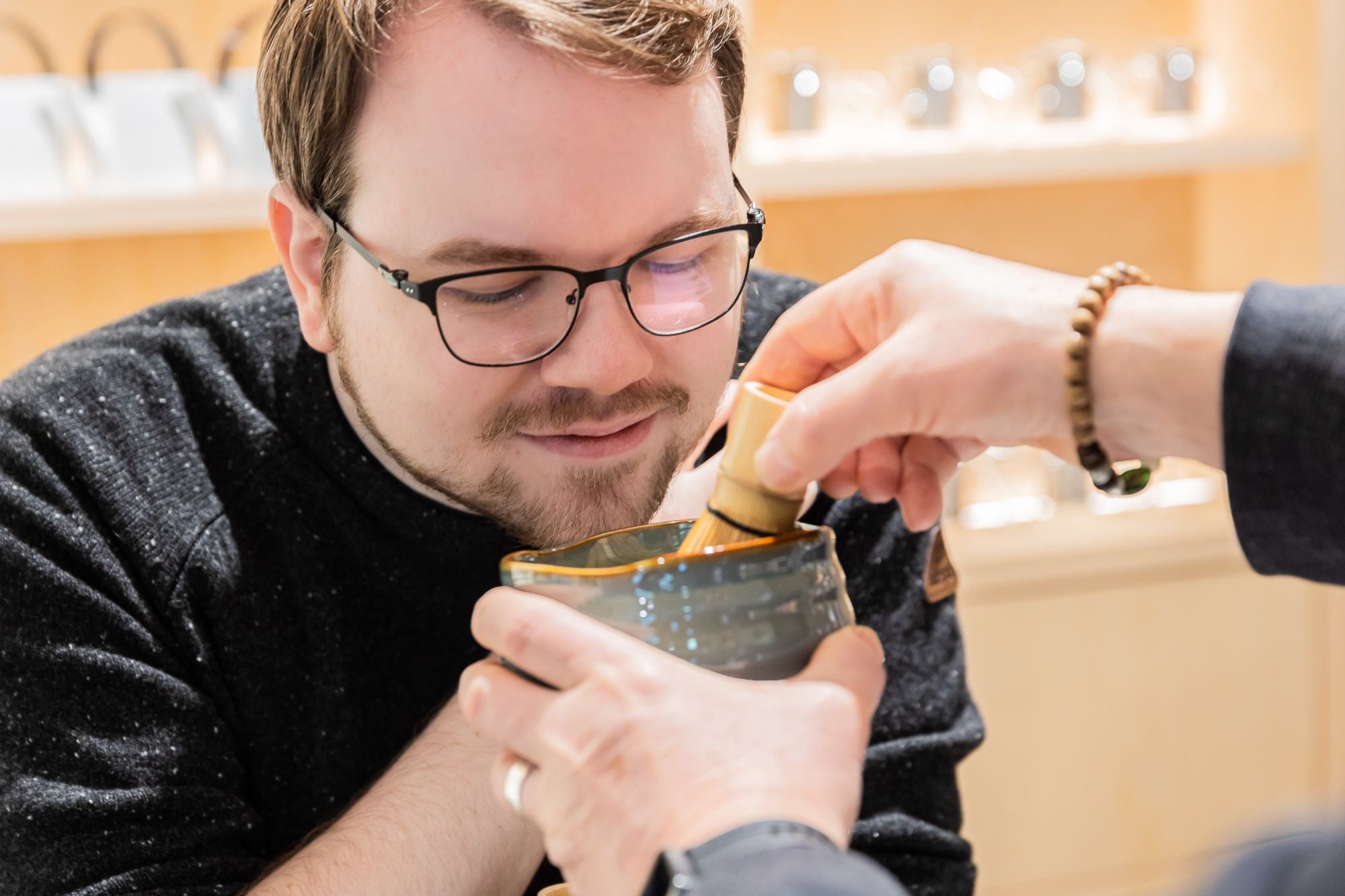 Mann mit Brille und Bart beobachtet, wie ein Bambusbesen Matcha in einer Keramikschale verquirlt wird, vor hellem Holzregal.