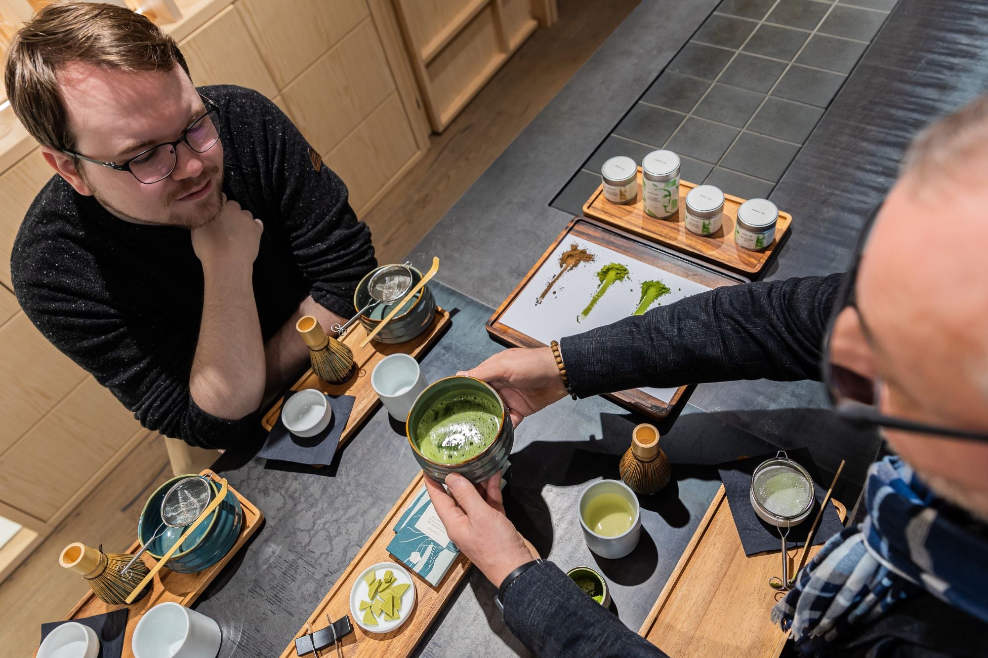 Zwei Männer konzentrieren sich bei einer Matcha-Tee-Zeremonie mit traditionellem Zubehör auf dunklem Holztisch