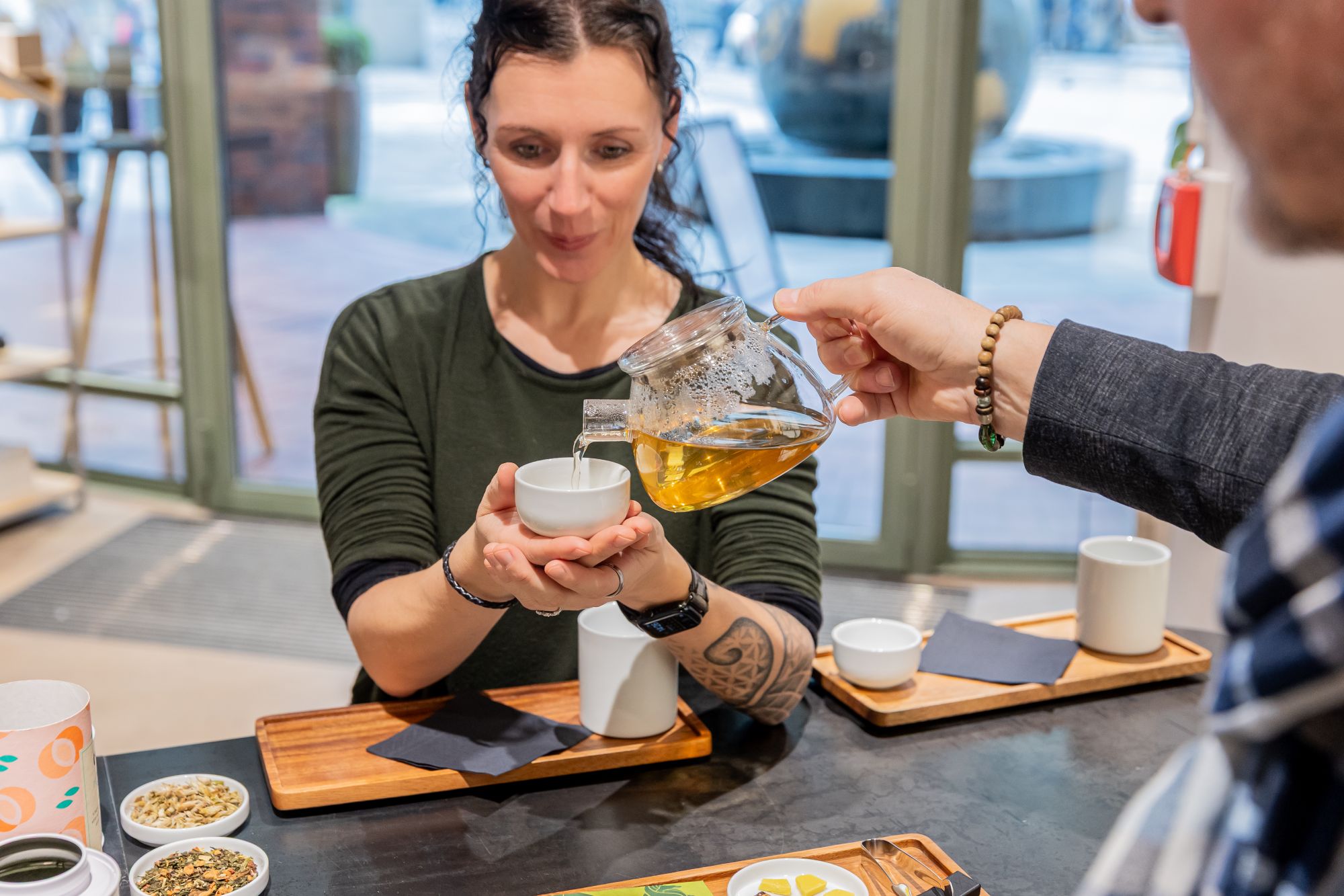 Frau in grünem Shirt hält eine Teetasse, Tee wird aus Glas-Teekessel eingegossen, bei Teezeremonie.