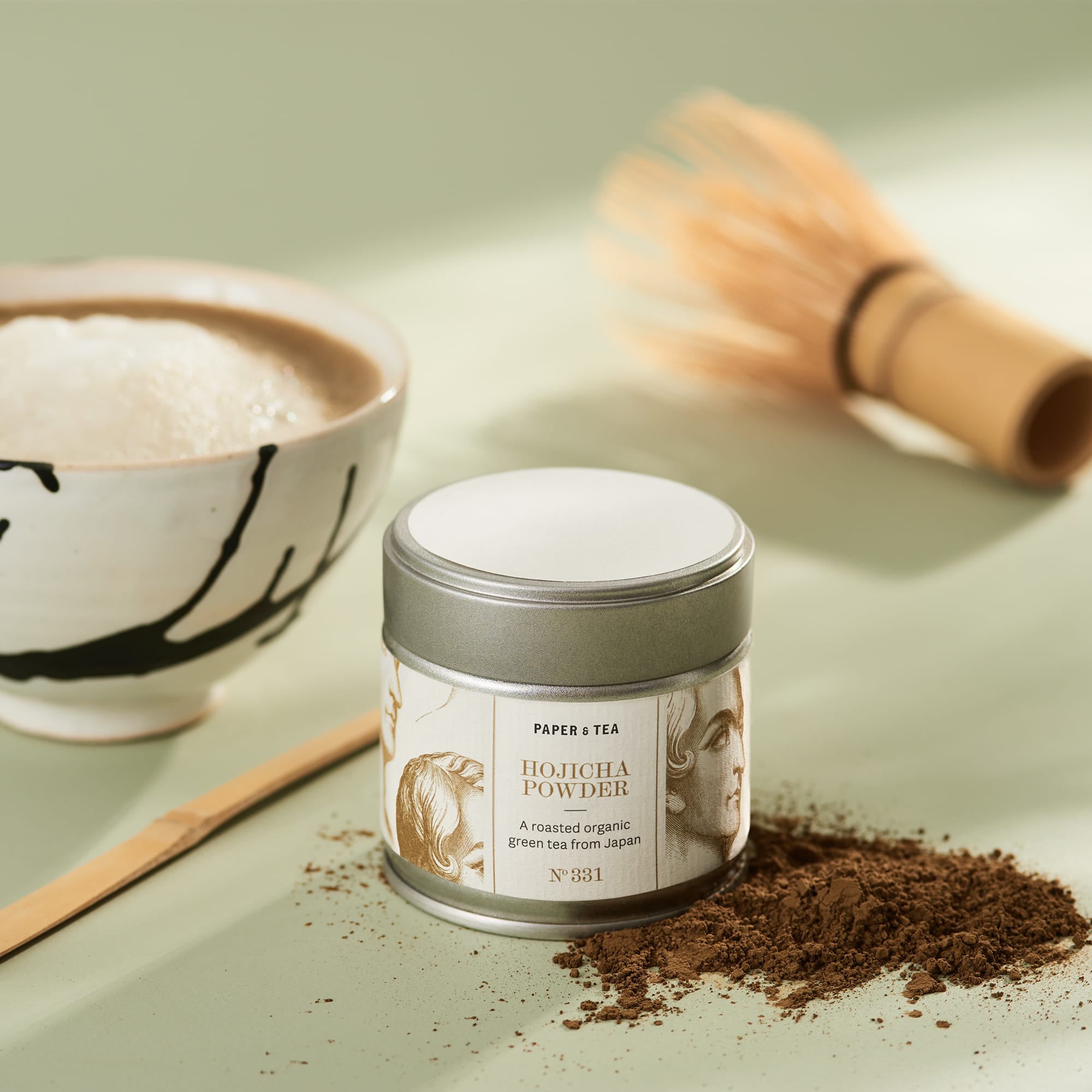 Bio Hojicha-Pulver in Metalldose, Keramikschale mit schaumigem Tee und Bambusbesen auf grünem Hintergrund