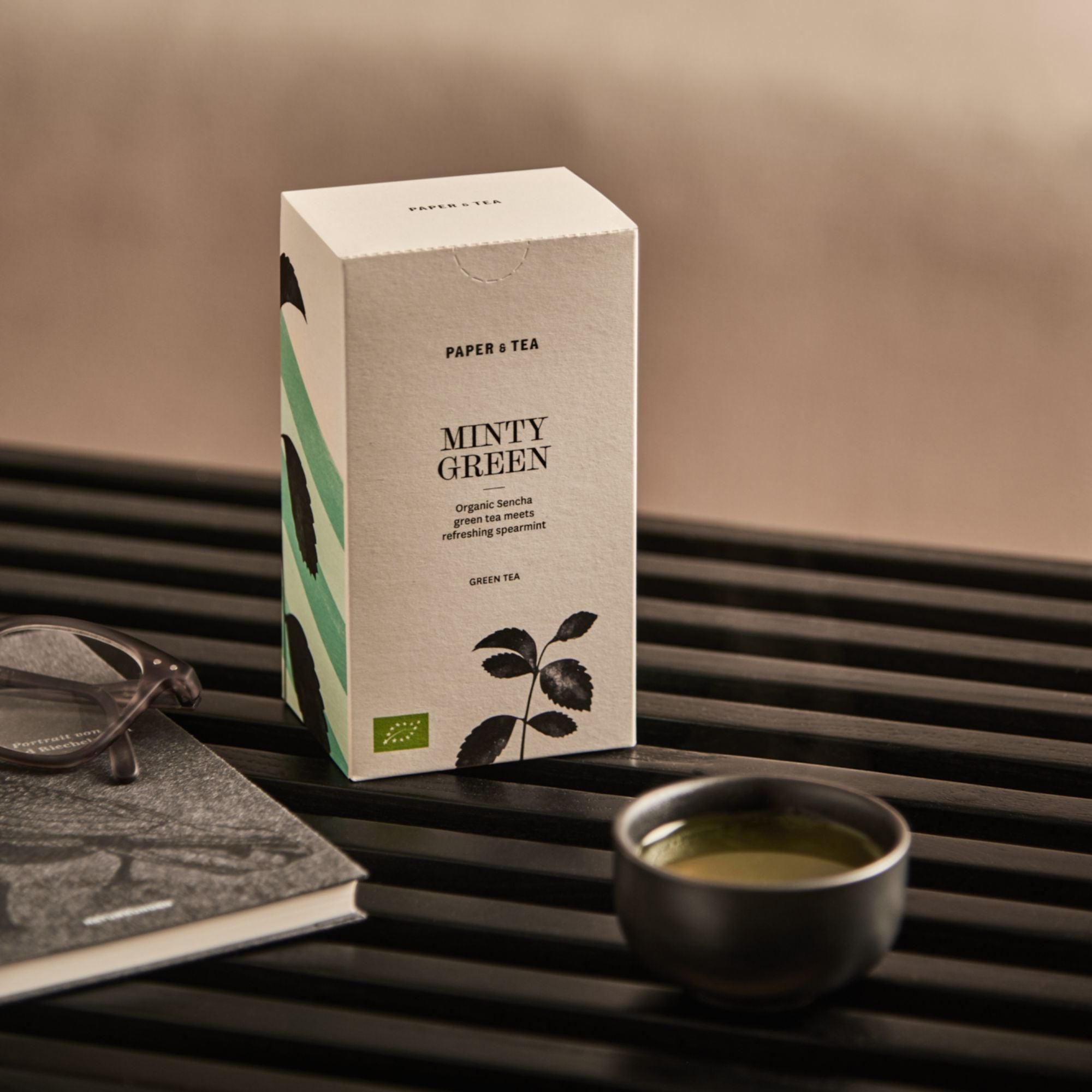 Packung PAPER & TEA Minty Green mit Bio-Sencha und Minze, daneben grüne Teetasse und Buch mit Brille auf Tisch.