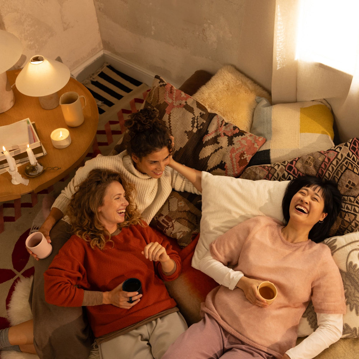 Drei lachende Frauen auf einem Sofa mit gemusterten Kissen, trinken Tee in gemütlicher Wohnzimmeratmosphäre.