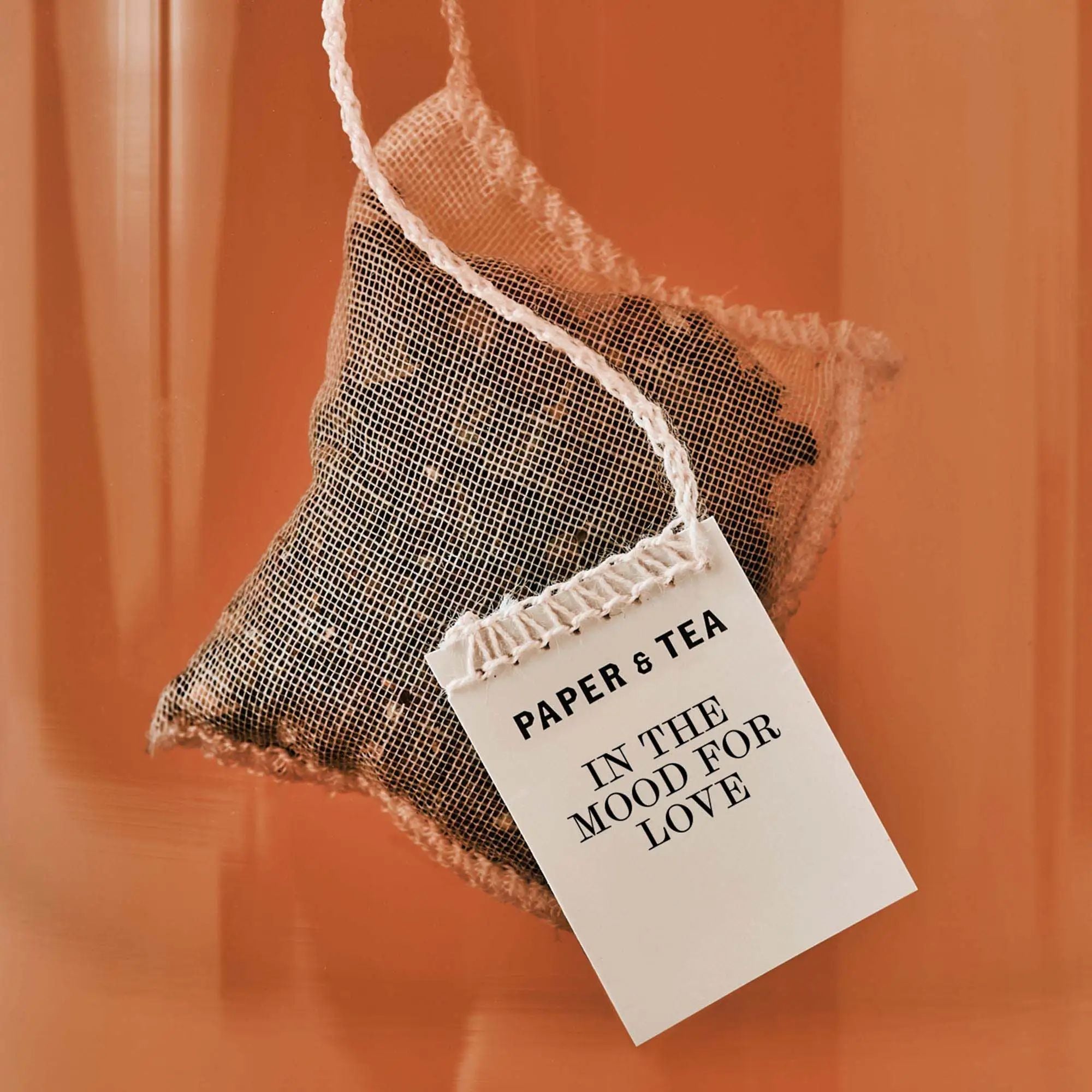 Feiner Mesh-Teebeutel mit losen Blättern und Anhänger 'PAPER & TEA IN THE MOOD FOR LOVE' vor orangem Hintergrund.