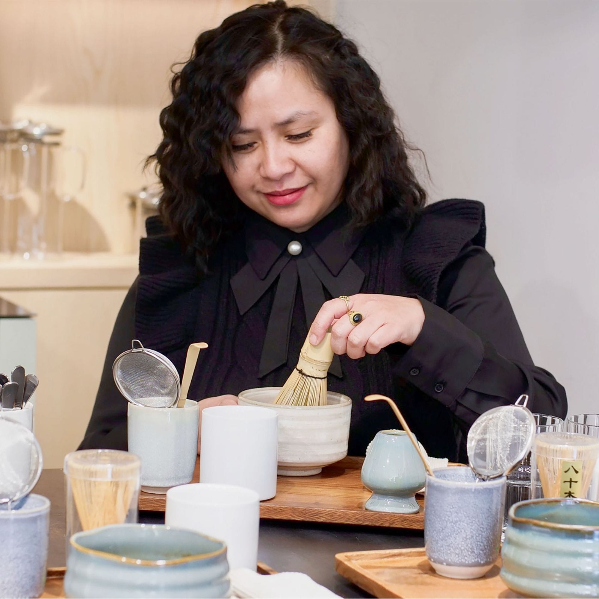 Frau mit lockigen dunklen Haaren bereitet Tee in einer Keramikschale mit Bambusbesen zu.
