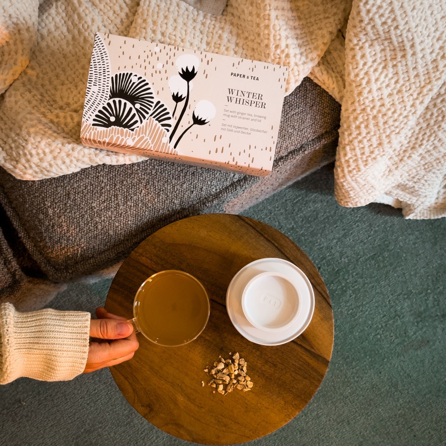 Rechteckige PAPER & TEA Winter Whisper Box auf Decke mit Glasbecher Ingwertee und getrocknetem Ingwer auf Holztisch.