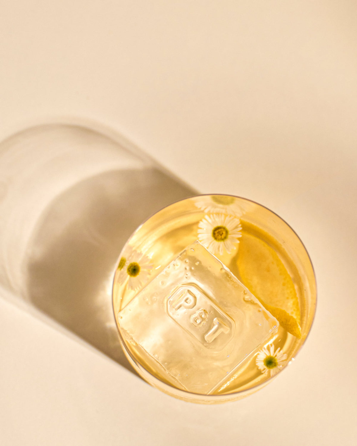 Glas mit hellgelbem Getränk, großem Eiswürfel mit P&T-Gravur, Gänseblümchen und Zitronenschale auf beigem Hintergrund.