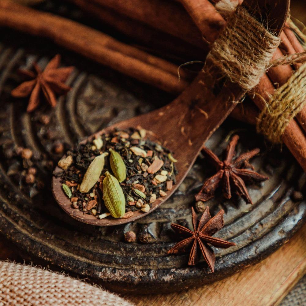 Holzlöffel mit Gewürzmischung aus grünem Kardamom und schwarzem Pfeffer auf dunklem Tablett mit Sternanis und Zimtstangen