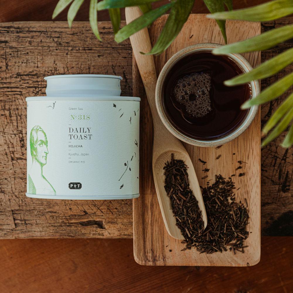 Weißer Teebehälter mit grünen Illustrationen, beschriftet mit HOJICHA Grüntee, neben Holztablett mit losem Tee.