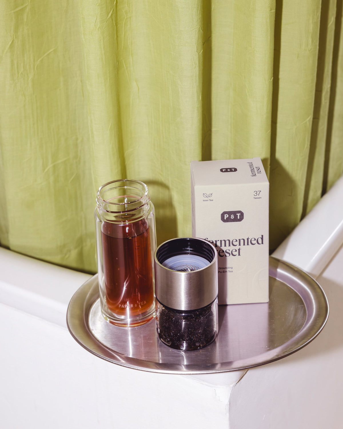 Transparente Flasche mit rotem Tee neben P&T Grounding Pu-Erh Teebox und Metallsieb auf rundem Tablett vor gelb-grünem Vorhang.