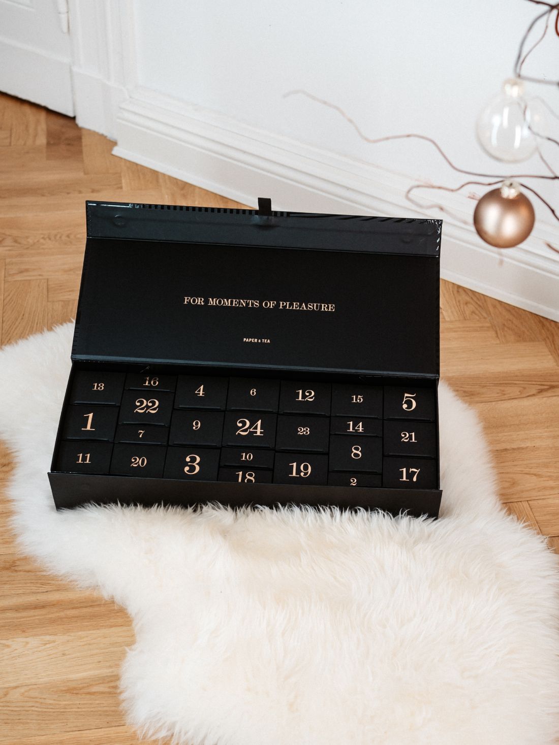 Schwarzer Paper & Tea Adventskalender mit goldenen Zahlen auf Fellteppich vor weißer Wand.