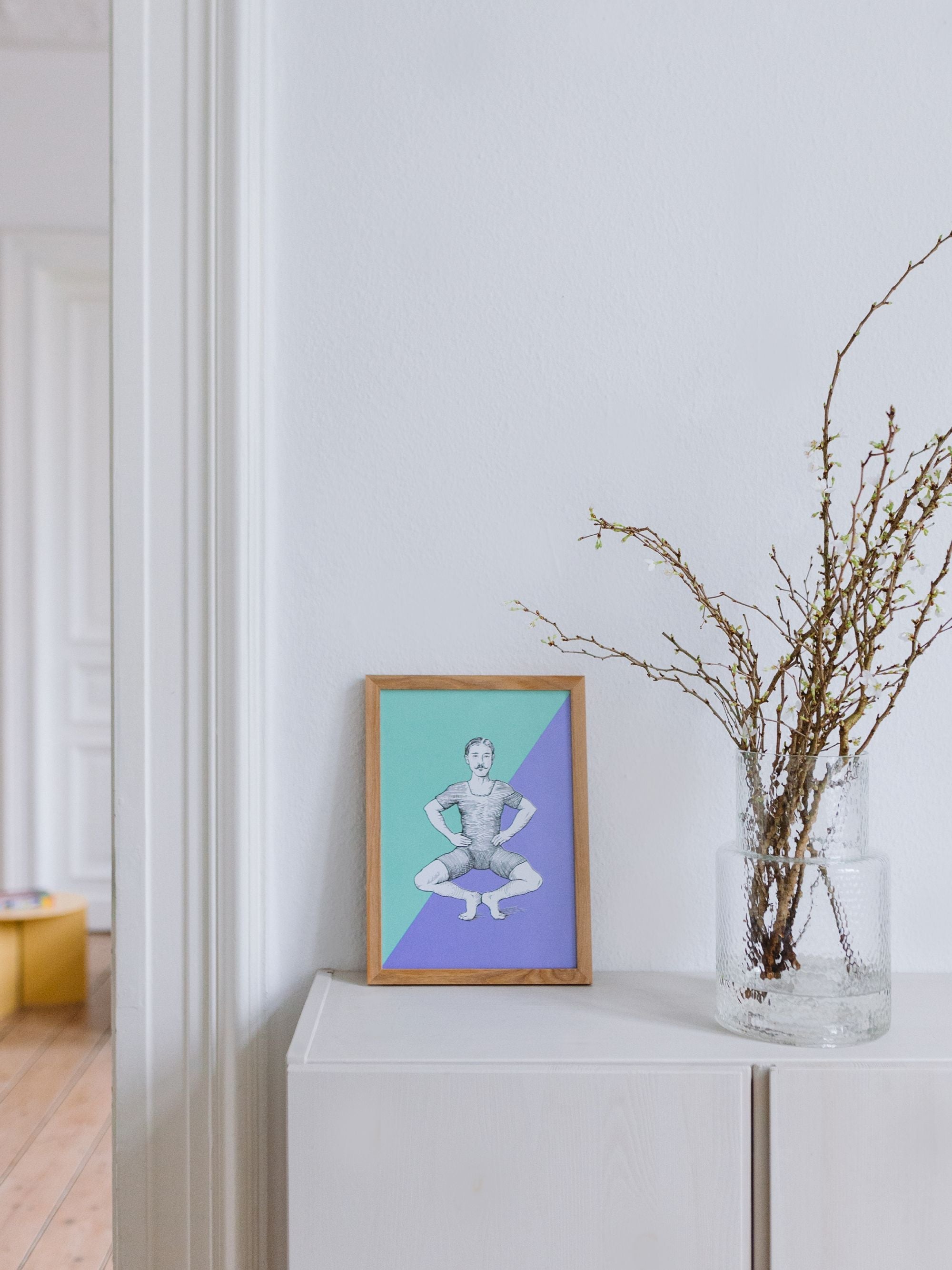 Minimalistisches Interieur mit weißem Sideboard, gerahmter Skizze einer hockenden Person und Glasvase mit Zweigen.