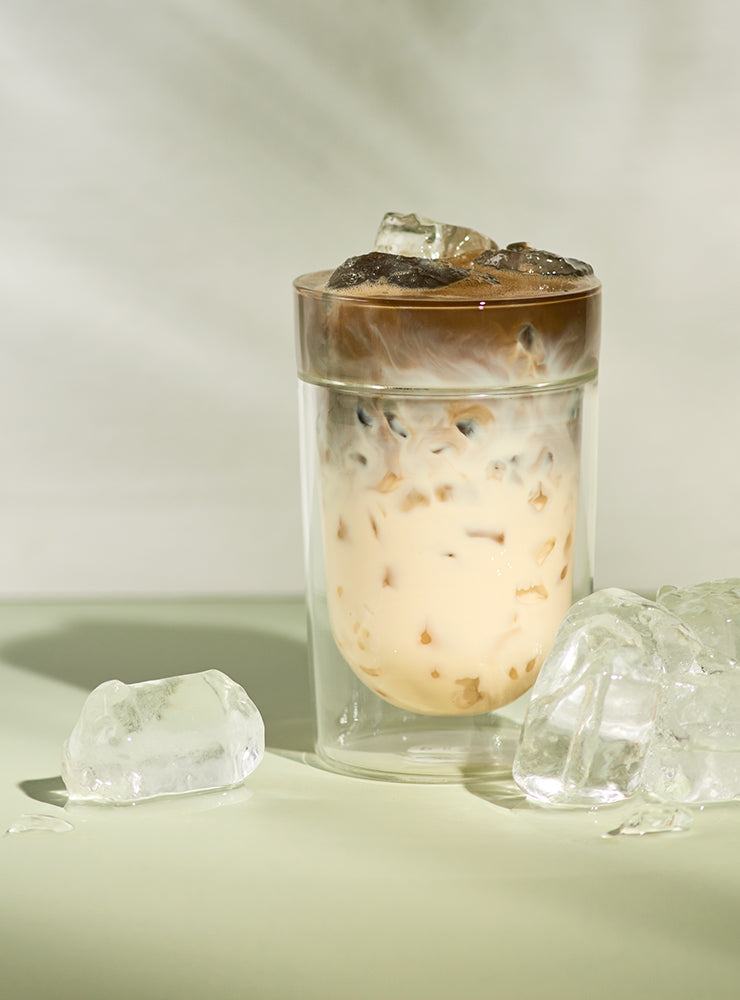 Klares Glas mit Eiskaffee und Milchwirbel, umgeben von schmelzenden Eiswürfeln auf hellem Untergrund.