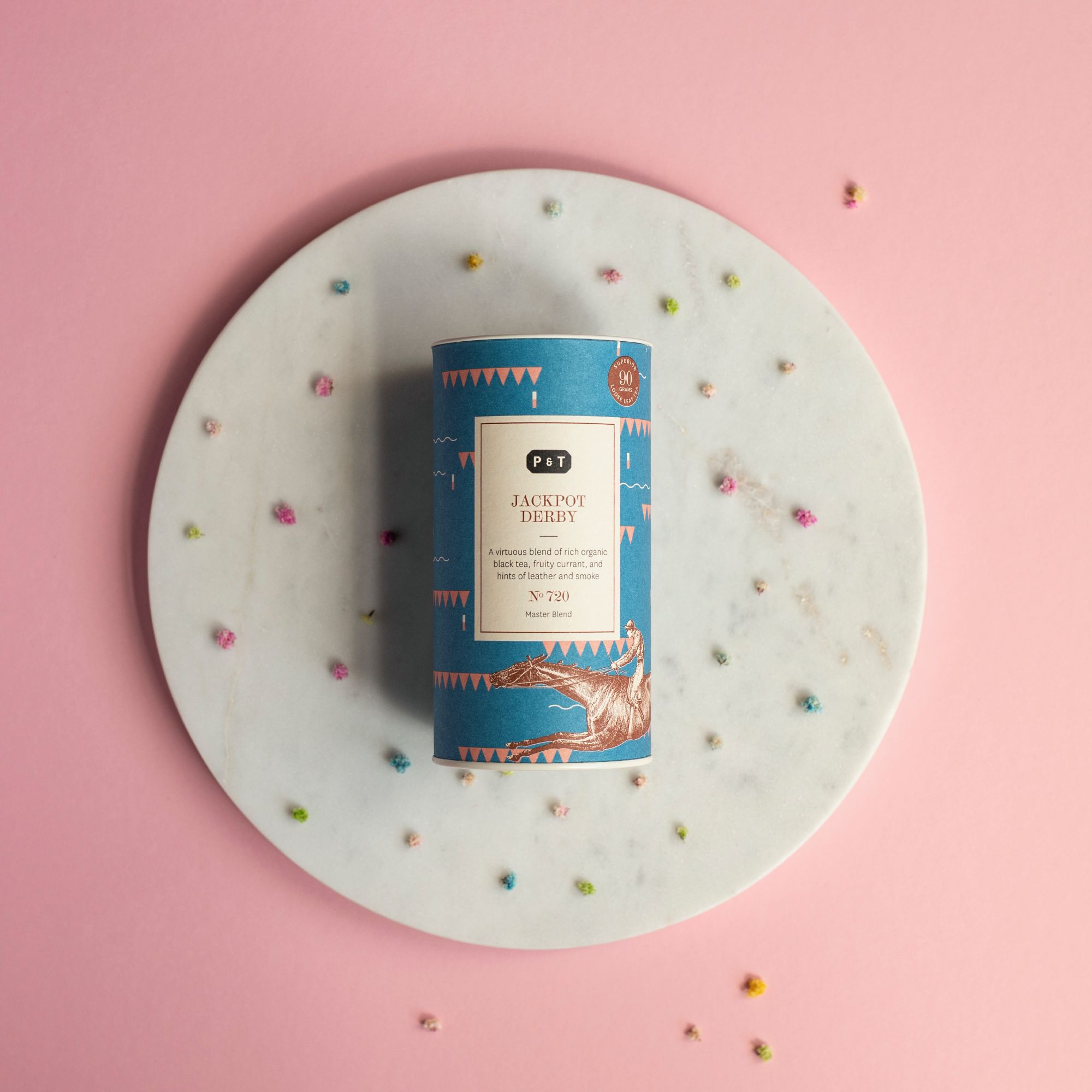 Zylindrische Teedose mit blauem Etikett und Reiter-Motiv auf weißem Marmorteller, umgeben von bunten Blüten.