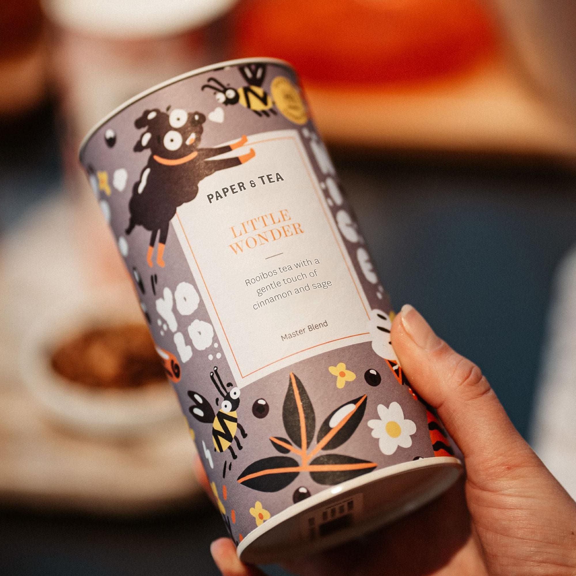 Hand hält bunte PAPER & TEA LITTLE WONDER Dose mit Rooibos-Tee, Zimt und Salbei, vor Teller mit Keksen.