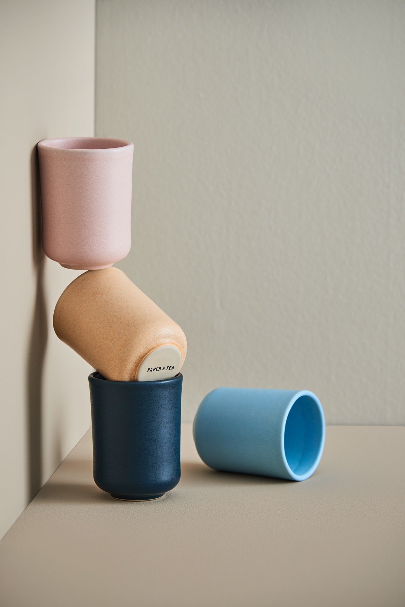 Vier pastellfarbene, mattierte Keramiktassen in Rosé-, Beige- und Blautönen, kunstvoll gestapelt, PAPER & TEA Schriftzug.