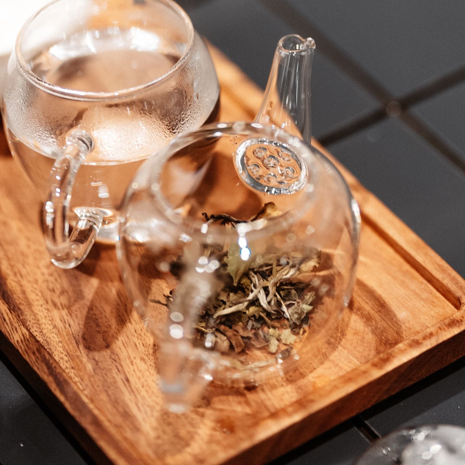 Transparente Glasteekanne mit getrockneten Teeblättern und Glas Wasser auf braunem Holztablett vor dunklen Fliesen.