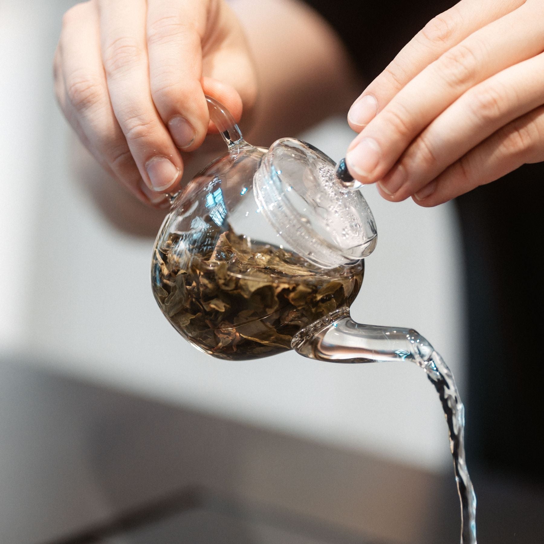 Zwei Hände gießen Tee aus einer transparenten Teekanne mit Teeblättern in ruhiger Atmosphäre.
