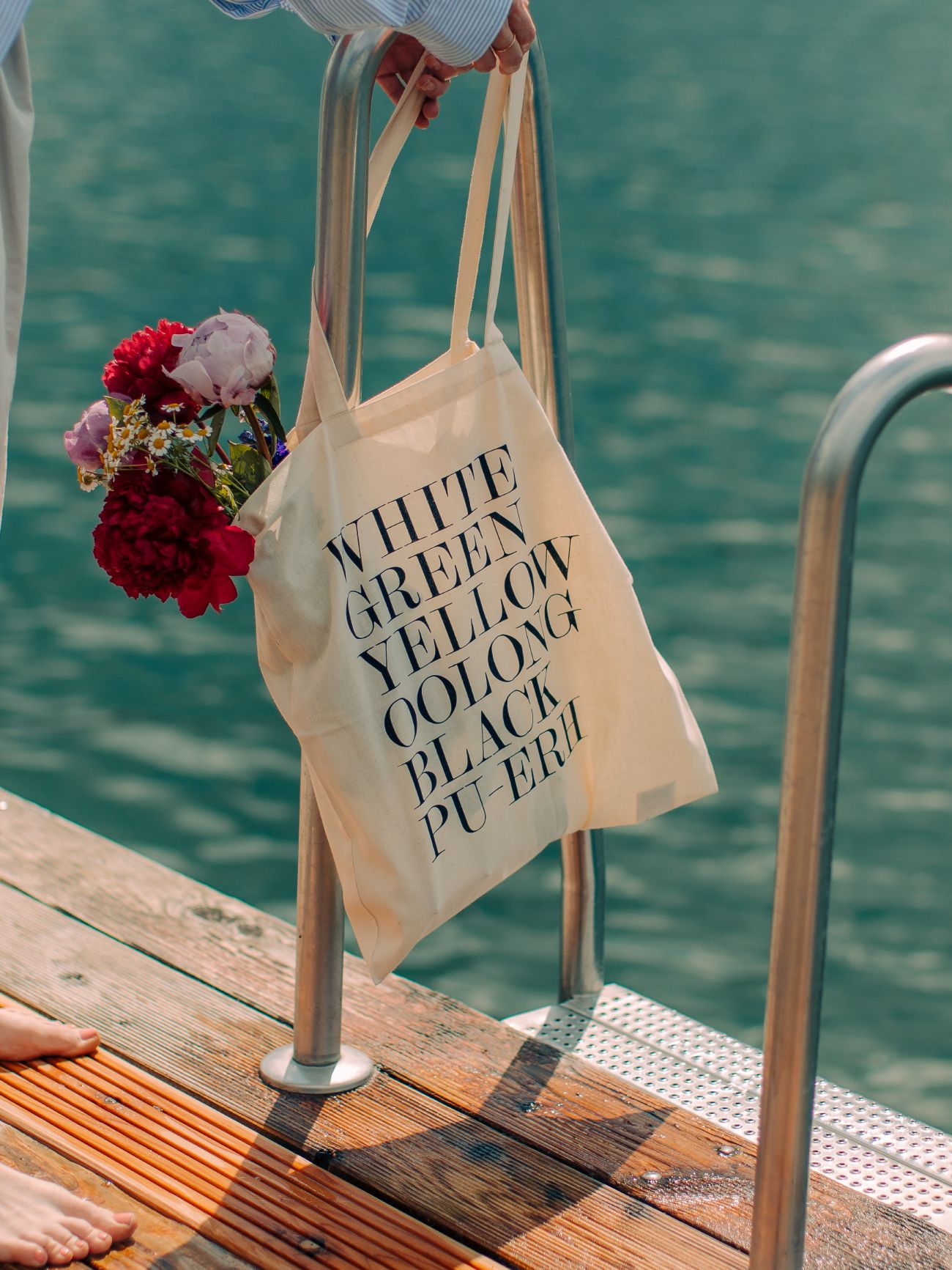 Cremefarbene Stofftasche mit Teeaufdruck und buntem Blumenstrauß, hängend an Metallleiter über ruhigem Wasser.