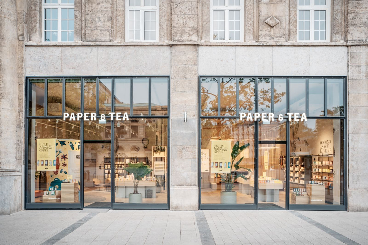 Moderner PAPER & TEA Teeladen mit großen Schaufenstern und Oolong-Tee-Werbung in historischem Gebäude.