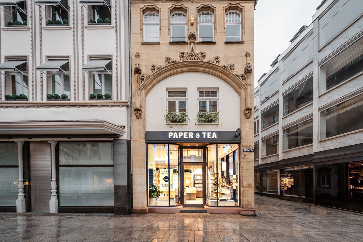 Kleines Schaufenster des PAPER & TEA Ladens mit schwarzem Schild und weißen Buchstaben, Pflanzen über dem Eingang.