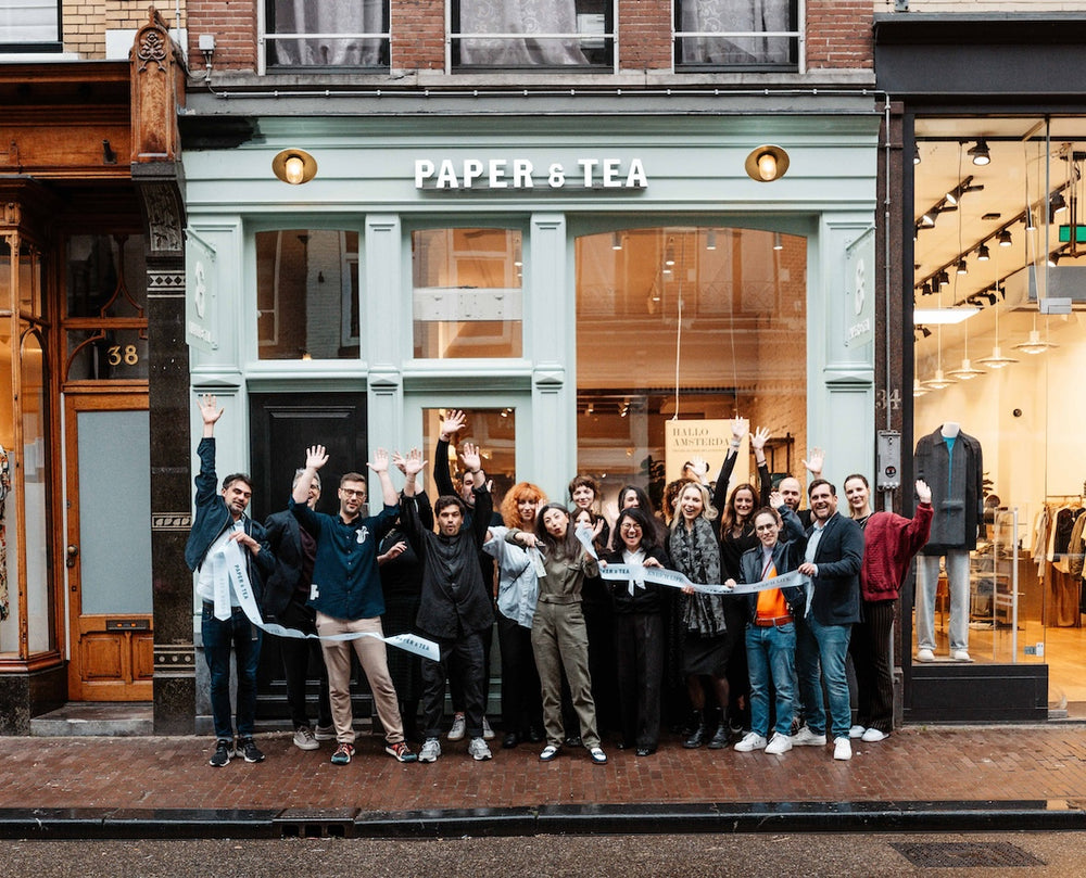 Fröhliche Gruppe vor mintgrünem PAPER & TEA Laden mit erhobenen Armen und Papierband, Hausnummern 38 und 34.