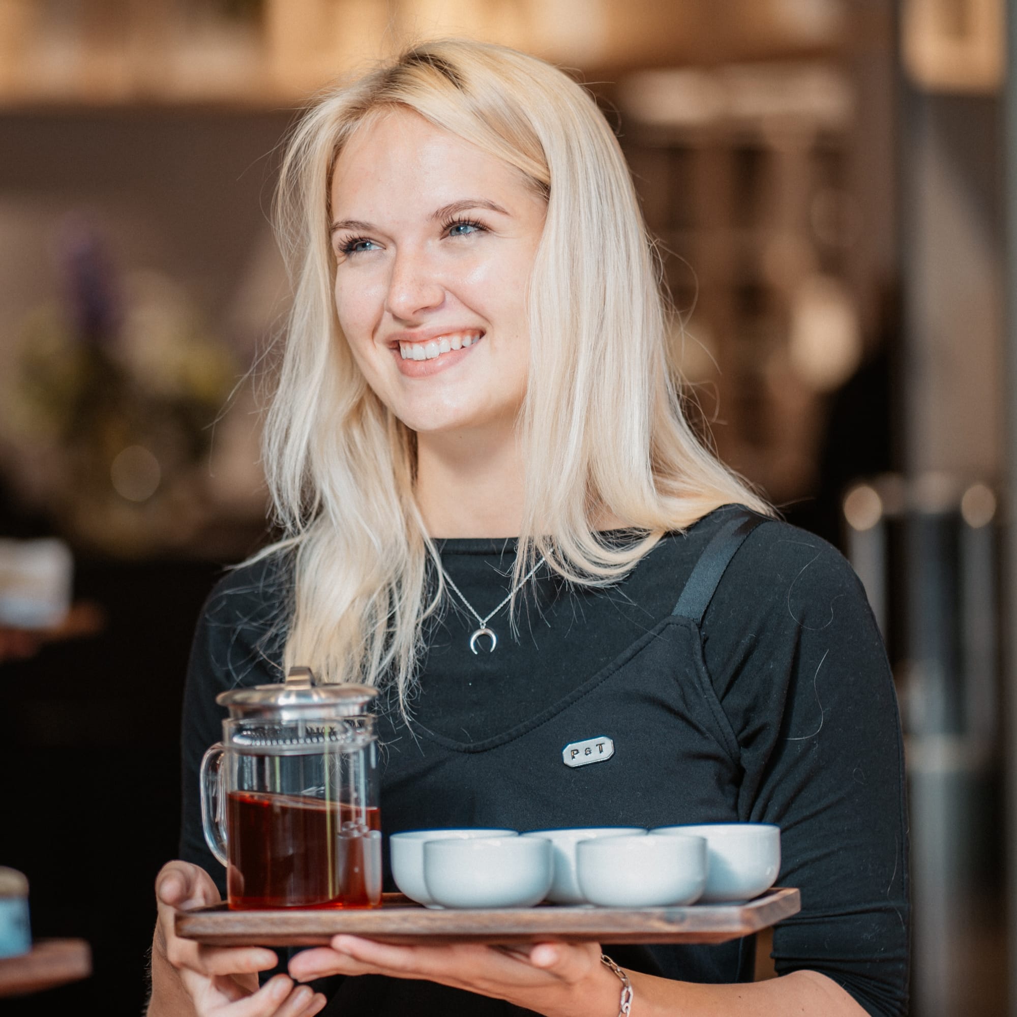 Lächelnde blonde Frau im schwarzen Oberteil serviert Tee im gemütlichen Café