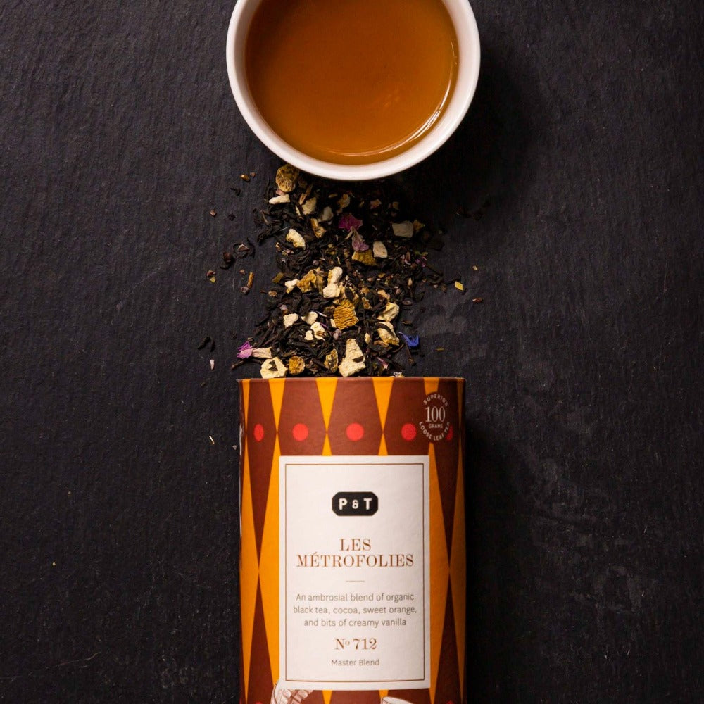 Zylinderförmiger Tee-Behälter LES MÉTROFOLIES mit Bio-Schwarztee, Orangenstücken, Vanille und Blüten, daneben Tasse Tee.