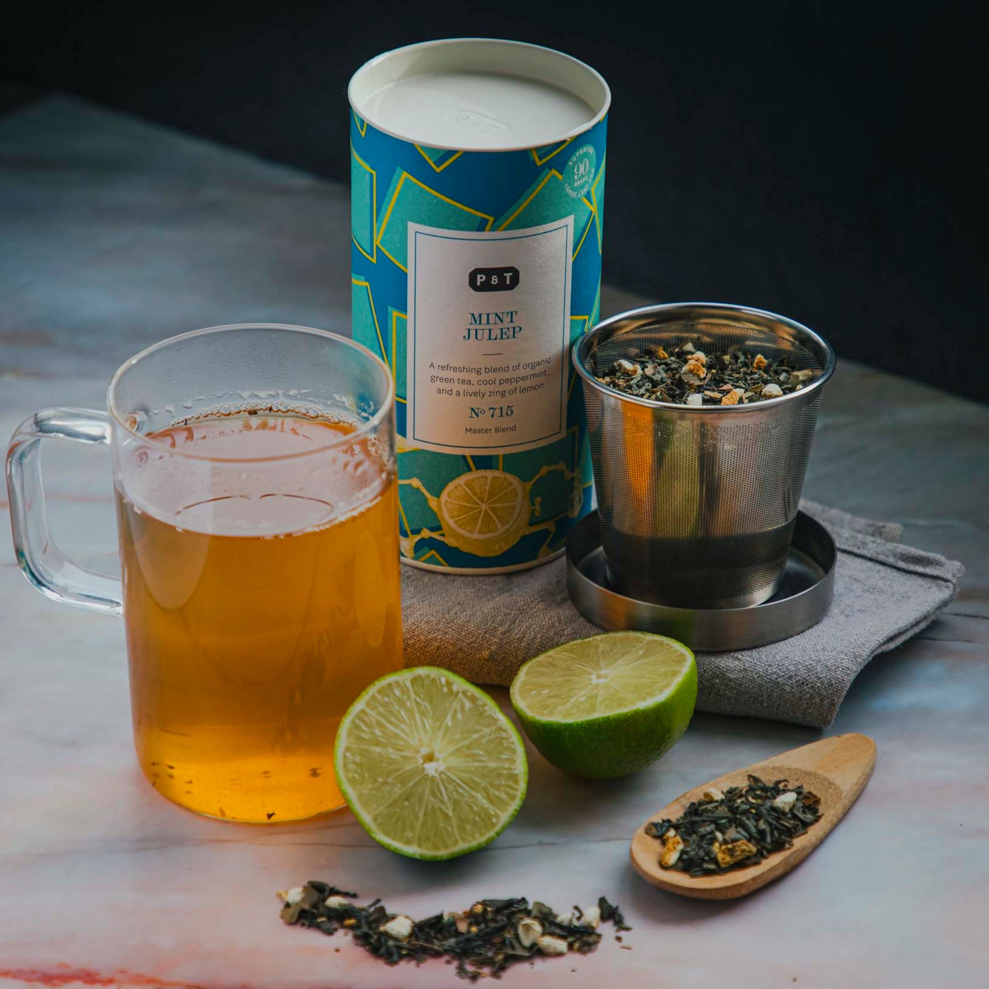 Blaue P & T Mint Julep Teedose mit Limettenhälften, Teeblättern, Teesieb und Glas grünem Tee auf Marmor.