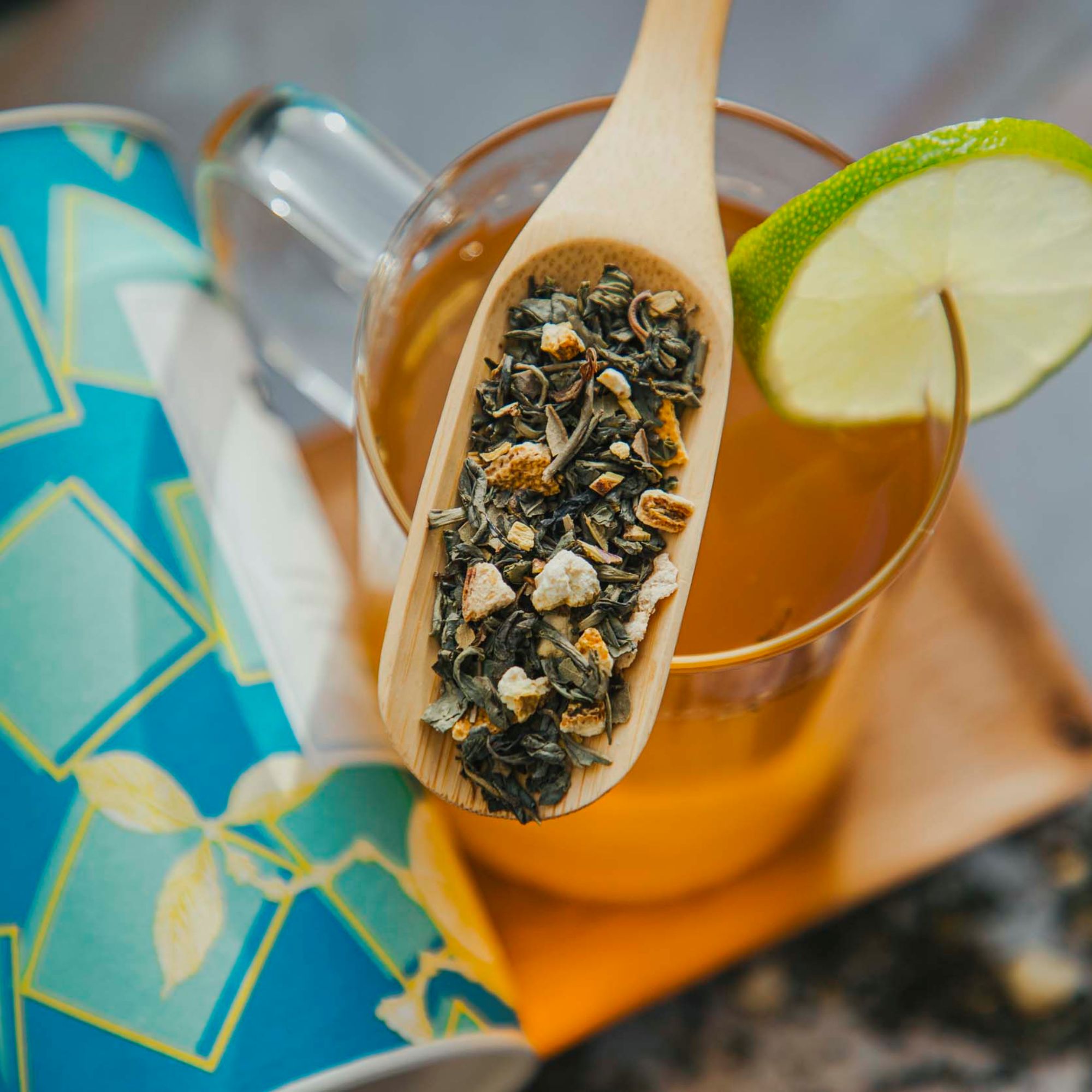 Ein Glas Tee mit Limettenscheibe, lose Teeblätter und Orangenschalen auf Holzlöffel, blaue Teedose.